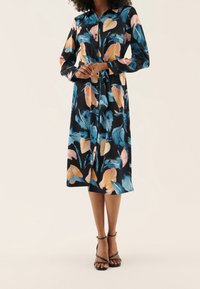 Mekko, jossa on kukkakuosi mustana, kevyt vaaleanpunaisten ja sinisten lehtien kanssa, pitkät hihat, kaulus ja vyötärölle sidottu vyö. Jalkineina on litteät sandaalit.