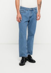 Blå denimjeans med rett benpassform, med fem lommer og glidelås, båret med svarte sko. Overflaten har en glatt tekstur.
