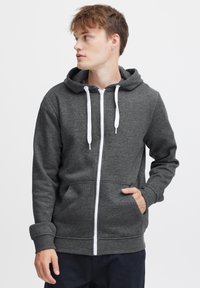 Sudadera con cremallera de color gris oscuro, fabricada en tejido suave, que cuenta con una capucha ajustable, cremallera blanca y bolsillos frontales. Textura suave y diseño casual.