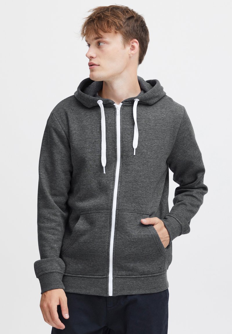 Sudadera con cremallera de color gris oscuro, fabricada en tejido suave, que cuenta con una capucha ajustable, cremallera blanca y bolsillos frontales. Textura suave y diseño casual.