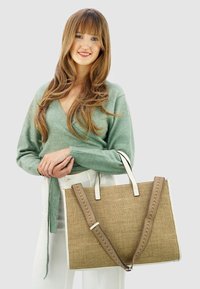 Grote beige shopper met een geweven textuur, witte handvatten en een patroonriem. Model in een groene trui en witte broek, houdend de tas vast.