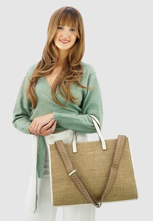 Grote beige shopper met een geweven textuur, witte handvatten en een patroonriem. Model in een groene trui en witte broek, houdend de tas vast.