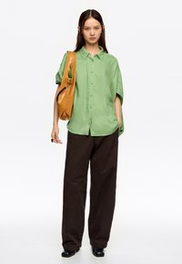 Camisa de botones verde claro, corte holgado, mangas cortas; pantalones anchos marrones; bolso de hombro de cuero marrón; botas negras.
