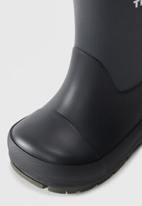 Bota de goma negra con punta redondeada, suela texturizada y acentos verdes en la base. Acabado suave con un detalle de costura en la parte superior.