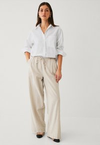Chemise blanche légère à manches retroussées, associée à un pantalon beige à cordon de serrage. Des mules noires complètent la tenue.
