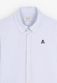 Camisa de cuadros azul claro hecha de tela con cuello abotonado. Presenta un pequeño logo de calavera en azul marino en el lado izquierdo del pecho y botones blancos.