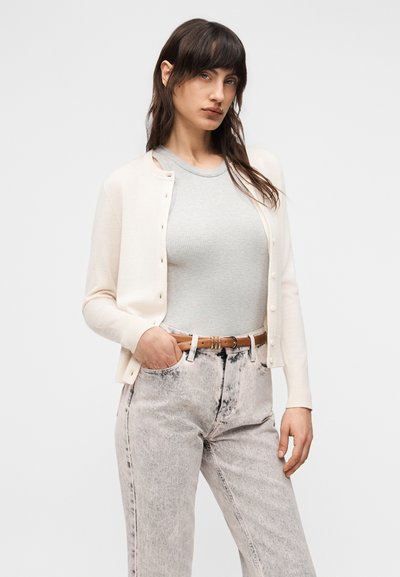 Γυναίκα με σκούρα μαλλιά που φοράει μπεζ ζακέτα πάνω από ανοιχτό γκρι τοπ χωρίς μανίκια, ξεβαμμένα γκρι τζιν ψηλόμεσα και καφέ ζώνη, ποζάροντας με το ένα χέρι στην τσέπη.