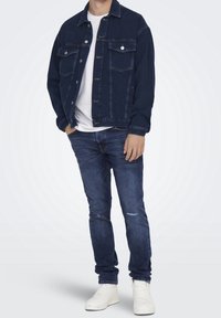 Mörkblå denimjacka med två bröstfickor, silverknappar och en avspänd passform, kombinerad med ljusblå slitna jeans och vita sneakers.