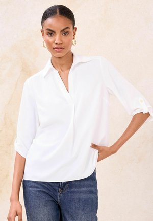 White, long-sleeve blouse with a v-neck, rolled cuffs, and a loose fit. Features a smooth texture and subtle draping, paired with dark blue jeans.