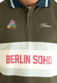 Oliven grøn polo shirt med broderet blomsterdesign, hvide og lyseblå vandrette striber, og "BERLIN SOHO" i fede røde bogstaver.