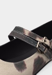 Κοντινό πλάνο ενός υφασμάτινου λουριού παπουτσιού με καφέ και μπεζ σχέδιο tie-dye και δύο χρυσούς μεταλλικούς ιμάντες πάνω από μαύρη σόλα.