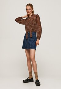 Blusa a maniche lunghe floreale marrone con un nodo in vita, abbinata a una minigonna di jeans blu con bottoni frontali. Mocassini neri e calzini marroni.