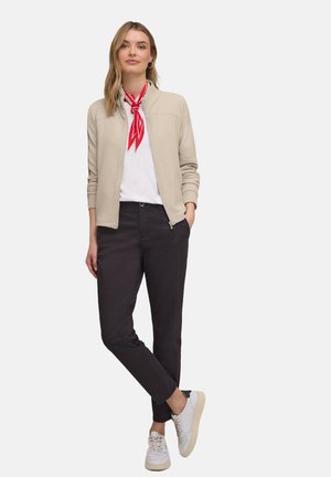 Femme portant une veste zippée texturée beige, une chemise blanche, un foulard rouge autour du cou, un pantalon noir et des baskets blanches, debout avec une main dans la poche.