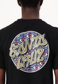 Tricou negru cu un grafic circular pe care se află textul "SANTA CRUZ" în aur, înconjurat de un design colorat cu motive de ochi, având accente de albastru și roz.