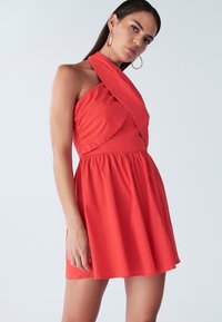 Červené šaty bez rukávů s řaseným výstřihem a přiléhavým živůtkem, s rozšířenou sukní, vyrobené z hladké látky.