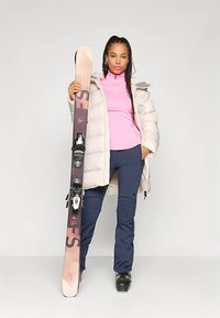 Bílý vatovaný kabát, růžové tričko s dlouhým rukávem, tmavě modré lyžařské kalhoty a černé lyžařské boty. Drží hnědé a růžové lyže s vázáním. Jednoduché pozadí.