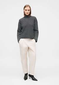 Μπλούζα ζιβάγκο με ασπρόμαυρες ρίγες συνδυασμένη με ανοιχτό μπεζ παντελόνι και μαύρα μυτερά μποτάκια αστραγάλου, στέκεται σε ουδέτερο φόντο.