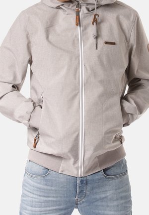 Jasnoszara kurtka na suwak z kapturem i sznurkiem, z prążkowanymi mankietami, noszona na jasnoniebieskich dżinsach, ręce wsunięte w przednie kieszenie.