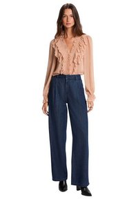 Mujer con blusa rosa claro con volantes, vaqueros de pierna ancha y talle alto azul oscuro, y botines negros de tacón, de pie sobre un fondo blanco.