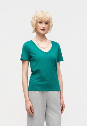 Młoda osoba z krótkimi kręconymi blond włosami, ubrana w zieloną koszulkę z dekoltem w serek, szare spodnie oraz podwójny naszyjnik z pereł na jednolitym tle.