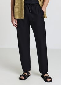 Schwarze Hosen mit lockerem Schnitt, elastischem Bund und seitlichen Taschen, kombiniert mit schwarzen Sandalen. Strick leichtes, helles Hemd.