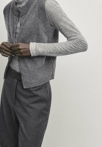 Person wearing layered grey knit cardigan over a light grey long-sleeve top and loose dark grey trousers against a plain background.