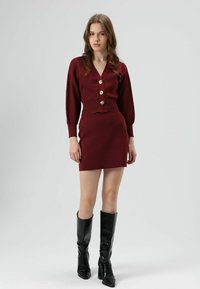 Cardigan en tricot bordeaux avec de gros boutons, associé à une jupe ajustée assortie. Le mannequin porte des bottes noires montantes jusqu'aux genoux. Texture lisse, design simple.