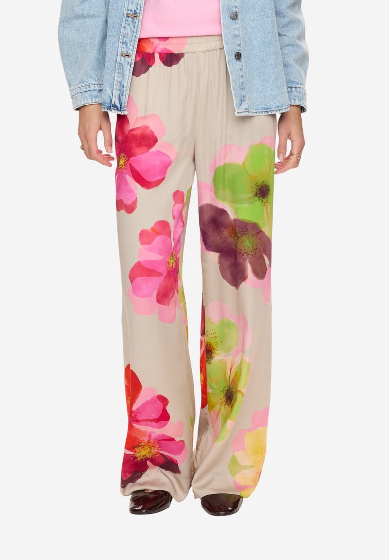 Femme portant un pantalon beige à jambes larges avec de grandes impressions florales colorées en rose, rouge, vert et violet, assorti à une veste en jean.