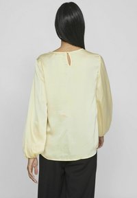 Nainen, jolla on pitkät tummat hiukset ja yllään vaaleankeltainen satiinipusero, jossa on pitkät puffihihat, ja mustat housut, seisoo yksinkertaista taustaa vasten.