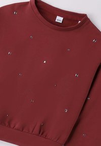 Felpa bordeaux con scollo rotondo, caratterizzata da borchie metalliche di forme varie sparse su tutto il tessuto e una fascia elastica in vita.