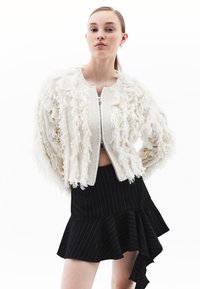 Giacca corta bianca texturizzata con dettagli a frange; presenta una chiusura con zip anteriore e polsini a costine. Indossata sopra una gonna asimmetrica nera a righe.