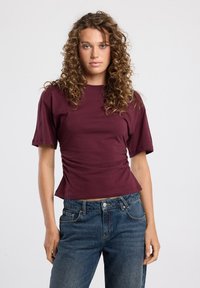 Burgundy short-sleeve top with a straight neckline and gathered side details, paired with high-waisted blue denim jeans. Smooth texture.