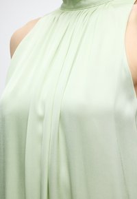 Αμάνικη ανοιχτό πράσινη μπλούζα με ψηλό ραμμένο λαιμό, λείο ύφασμα και διακριτικές κάθετες πιέτες στο εμπρός μέρος.