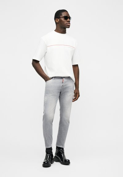 Homme portant une chemise blanche à manches courtes, un jean gris clair avec des déchirures discrètes, des bottines noires brillantes et des lunettes de soleil foncées, debout avec une main dans la poche.