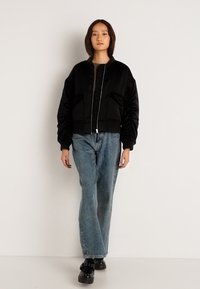 Μαύρο bomber jacket με ριγέ μανσέτες και γιακά, διαθέτοντας φερμουάρ και πλαϊνές τσέπες. Συνδυάζεται με ανοιχτό μπλε τζιν ίσιας γραμμής και μαύρα παπούτσια.