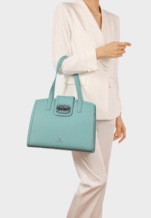 Donna in completo bianco che tiene una borsa strutturata color teal con doppie fibbie e una chiusura decorativa all'altezza della vita.