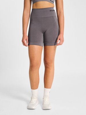 Femme portant un cuissard de cyclisme gris sans coutures taille haute, des chaussettes blanches et des chaussures de sport blanches, debout devant un fond clair uni.