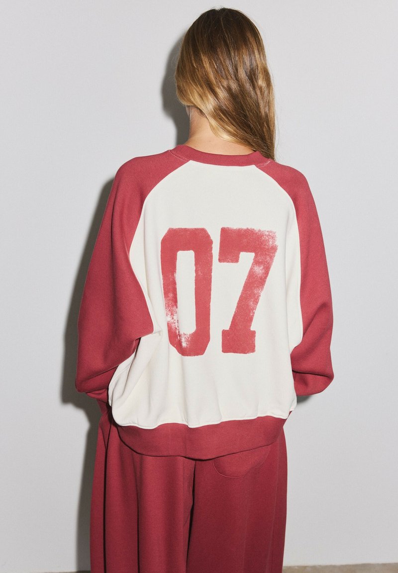Personne aux longs cheveux portant un sweat-shirt rouge et blanc avec le numéro 07 dans le dos et un pantalon rouge assorti, debout contre un mur uni.