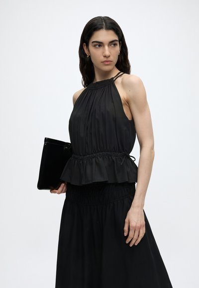 Γυναίκα που φοράει μαύρο αμάνικο τοπ halter και φούστα, κρατώντας μαύρο φάκελο, κοιτάζει στο πλάι μπροστά σε λευκό φόντο.