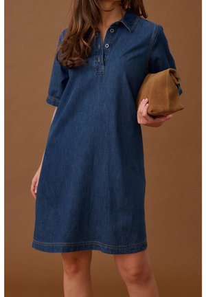 Femme portant une robe en jean bleue à manches courtes tenant une pochette en daim marron pliée devant un fond marron.