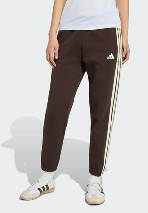 Personne portant un pantalon de survêtement Adidas marron avec des bandes blanches et un logo, des chaussettes blanches et des baskets blanches avec des bandes noires.