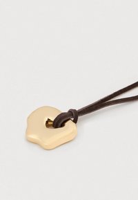 Κρεμαστό κόσμημα σε χρυσό τόνο με οργανικό σχήμα, προσαρτημένο σε καφέ δερμάτινο κορδόνι με λεπτομέρεια κόμπου. Λείο επιφάνεια χωρίς σχέδια.