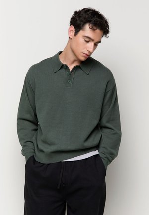 Jeune homme aux cheveux bouclés foncés, portant un pull polo vert à manches longues et un pantalon noir, debout les mains dans les poches, regardant vers le bas.