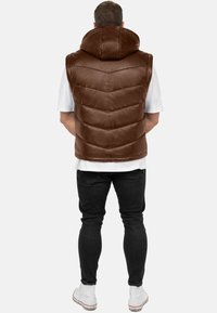 Gilet imbottito in pelle marrone con cappuccio alto, caratterizzato da cuciture trapuntate e una superficie liscia, indossato sopra una camicia bianca e jeans neri.