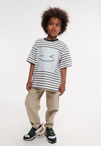 T-shirt in cotone a righe bianco e blu navy, con grafica di un volto sorridente; abbinato a pantaloni beige e sneaker multicolori.