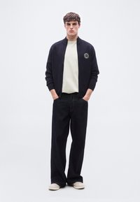 Νεαρός άνδρας στέκεται με τα χέρια στις τσέπες, φοράει μαύρο μπουφάν με φερμουάρ, λευκή μπλούζα, φαρδιά σκούρα τζιν και κρεμ αθλητικά παπούτσια, σε λευκό φόντο.