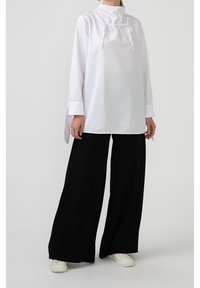 Witte lange mouw tuniek met een hoge kraag en trekkoorddetails, gecombineerd met brede zwarte broek. Schoenen zijn witte sneakers.
