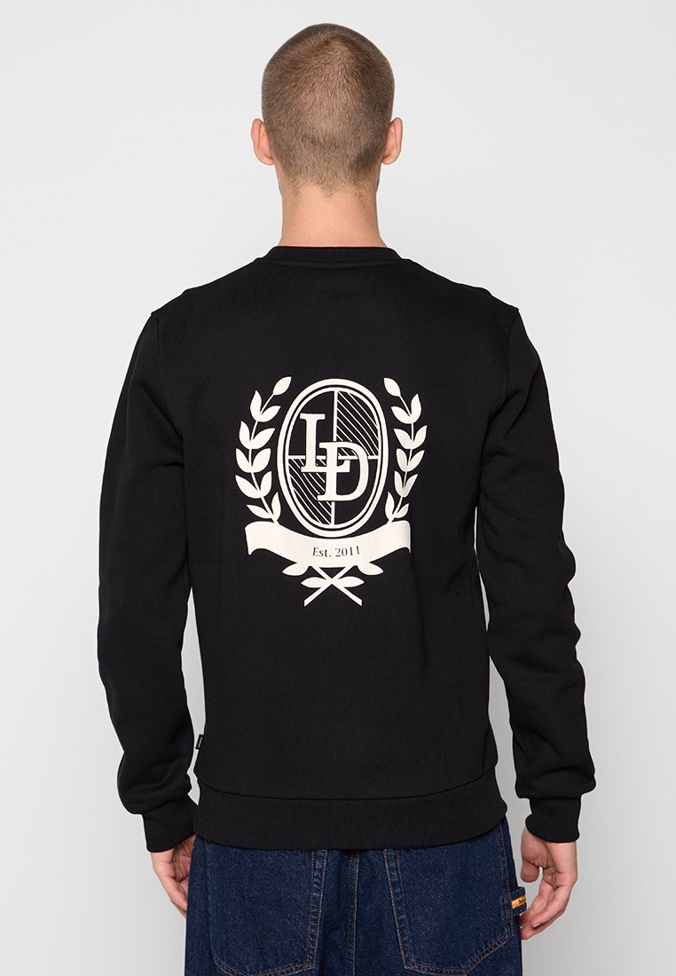 Mann trägt einen schwarzen Sweatshirt mit weißem LD-Logo und Blattkranz-Grafik auf dem Rücken, kombiniert mit blauen Jeans.