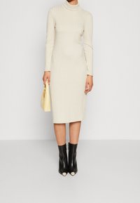 Robe en maille côtelée beige avec un col roulé, coupe ajustée. Associée à des bottines noires et un petit sac à main jaune.
