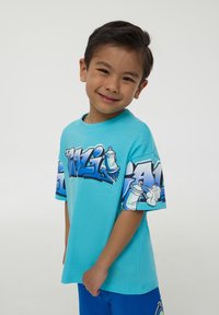 T-shirt turchese con grafica in stile graffiti che presenta bombolette di spray. Maniche corte, materiale in cotone morbido, vestibilità rilassata.